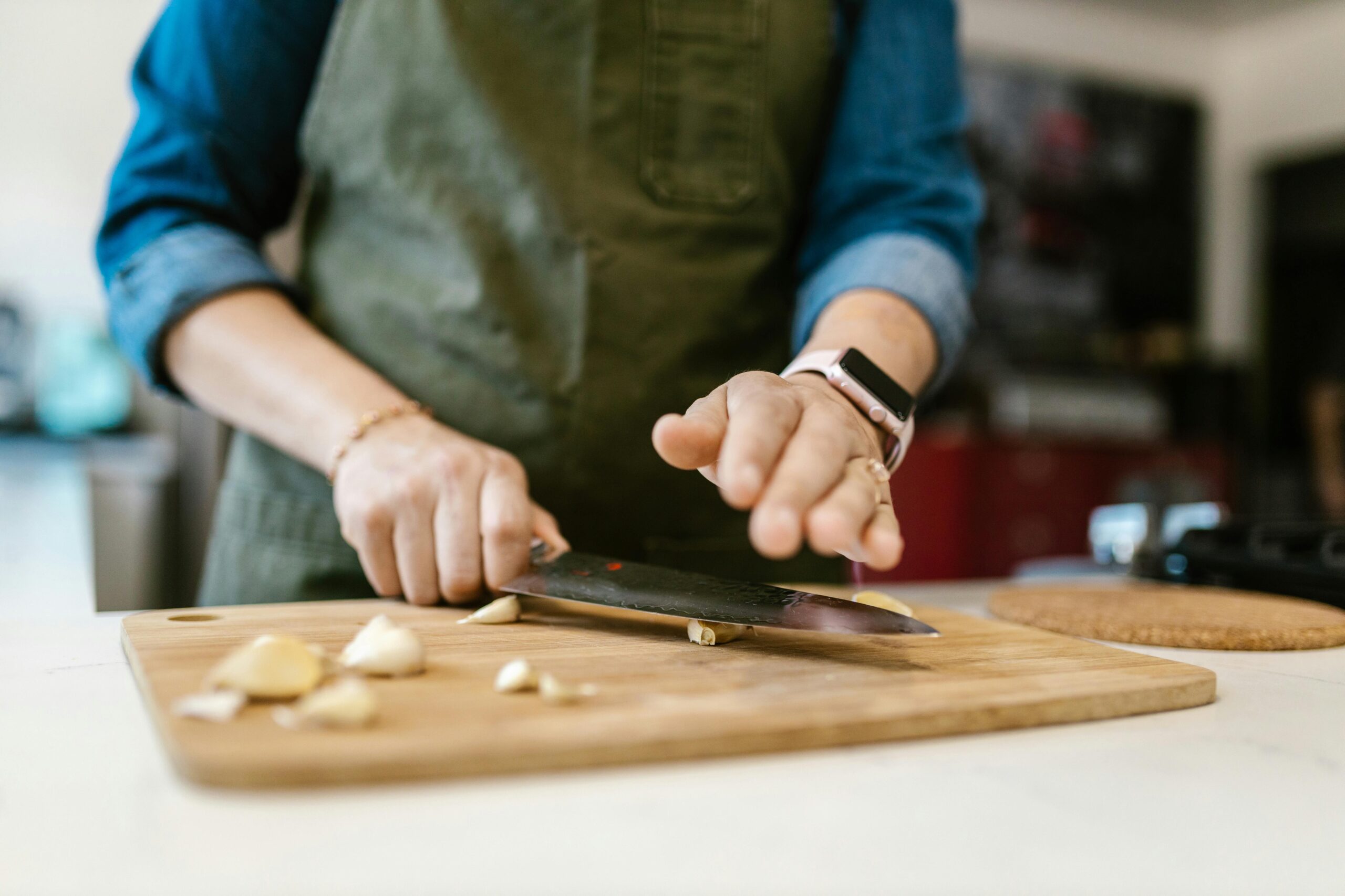 A person chops garlic on a wooden cutting board, with one hand guiding the knife while the other hand is raised for safety.