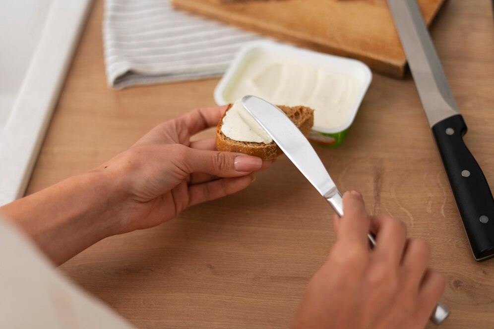 A person spreads cream cheese on a slice of whole grain bread with a knife, set on a wooden table with utensils nearby.