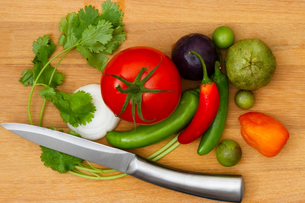Fresh vegetables and herbs including tomatoes, peppers, cilantro, and onions arranged beside a knife on a wooden cutting board.