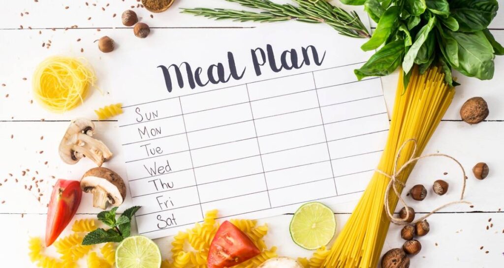 A meal plan sheet with days of the week, surrounded by fresh ingredients like pasta, tomatoes, mushrooms, lime, and herbs.