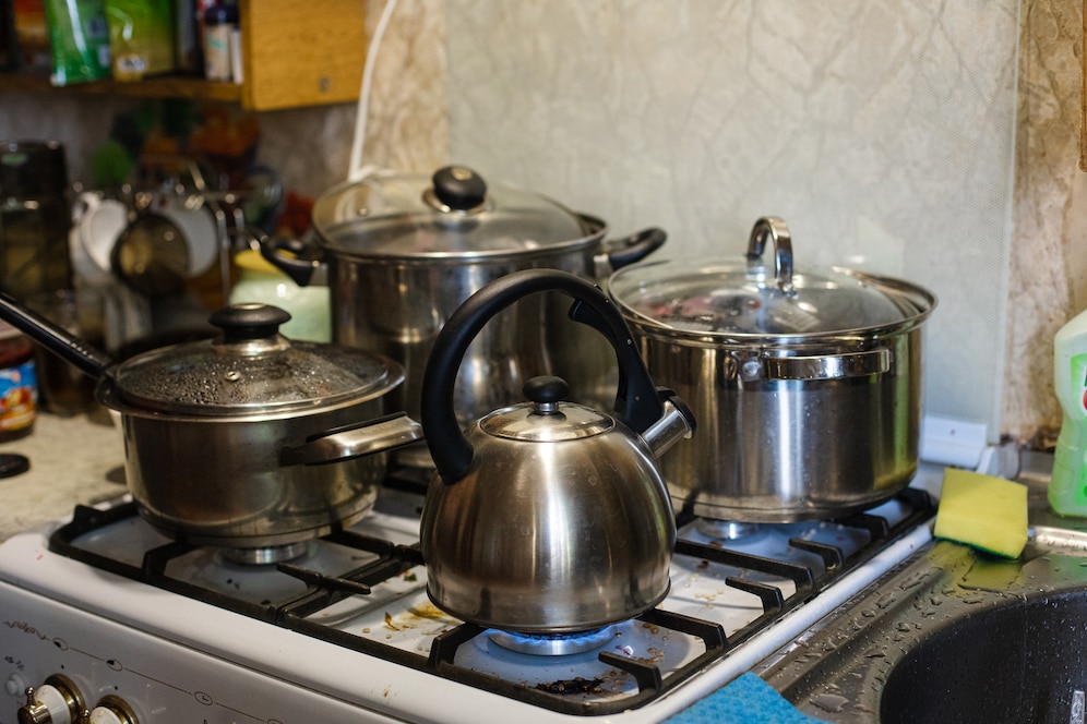 A cluttered stove with multiple stainless steel pots and a kettle, beside a sink with a sponge, in a busy kitchen setting.
