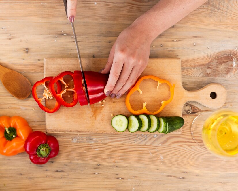 A hand slices a red bell pepper on a wooden cutting board, surrounded by colorful peppers, sliced cucumber, and a glass of yellow drink.