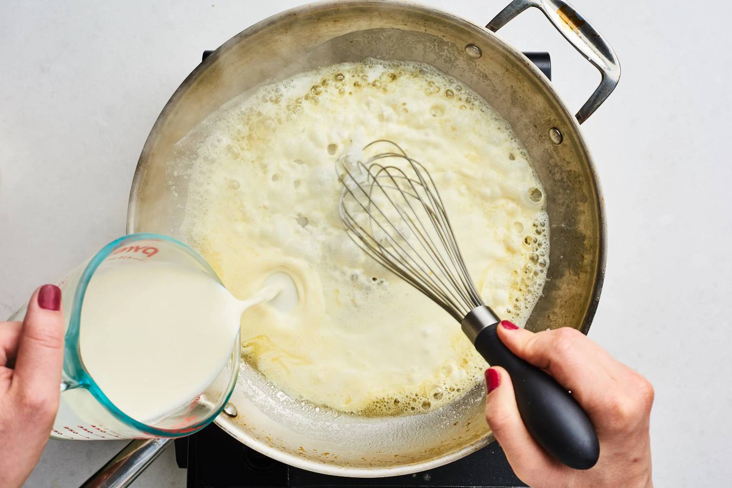 A hand pours milk into a bubbling saucepan, while the other hand whisk-stirs the frothy mixture. Steam rises from the pot.