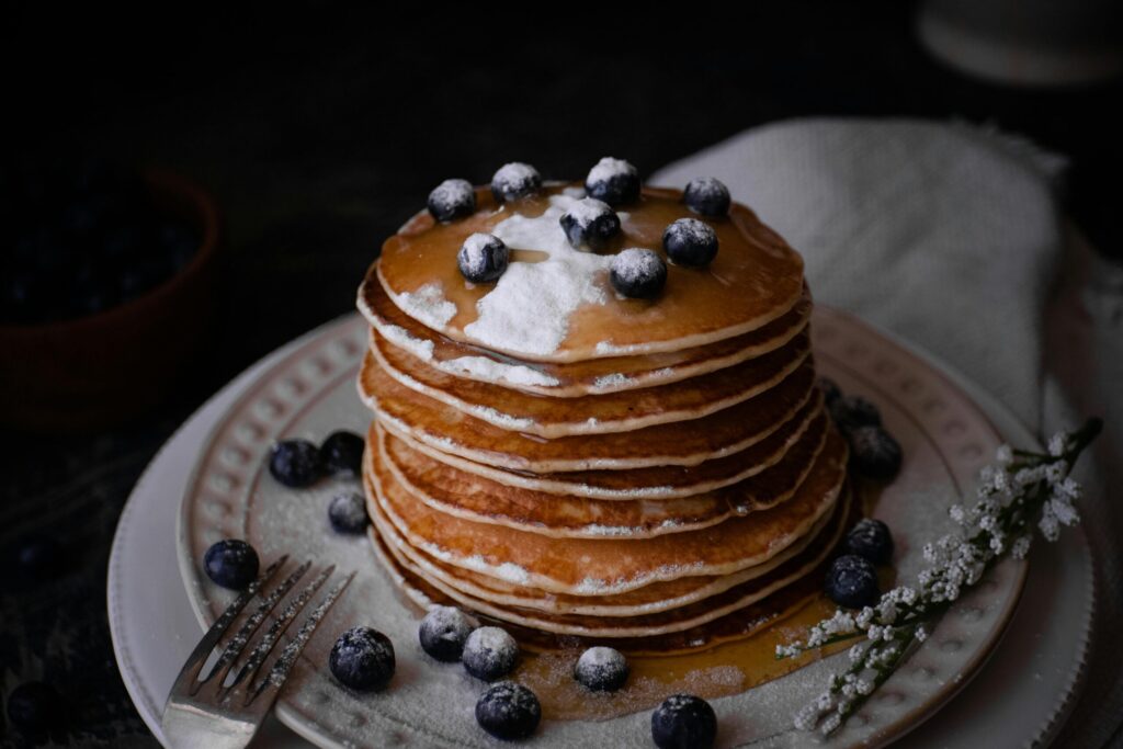 A stack of fluffy pancakes drizzled with syrup, topped with blueberries and powdered sugar, surrounded by fresh blueberries and a floral garnish.