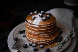 A stack of fluffy pancakes drizzled with syrup, topped with blueberries and powdered sugar, surrounded by fresh blueberries and a floral garnish.