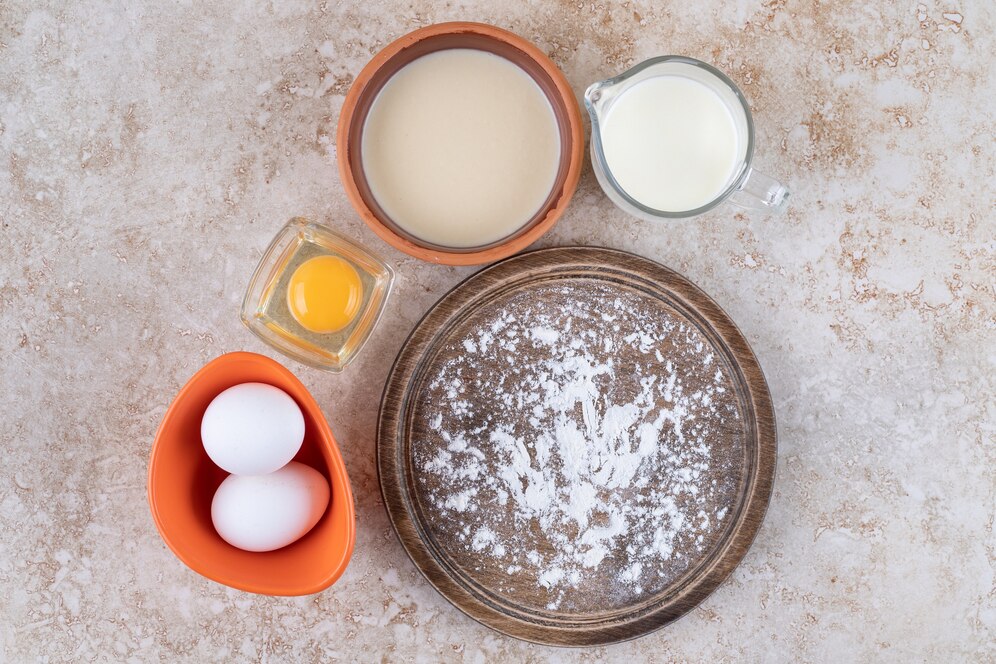 Ingredients for baking laid out: eggs, milk, flour, and a bowl of cream on a marble countertop.