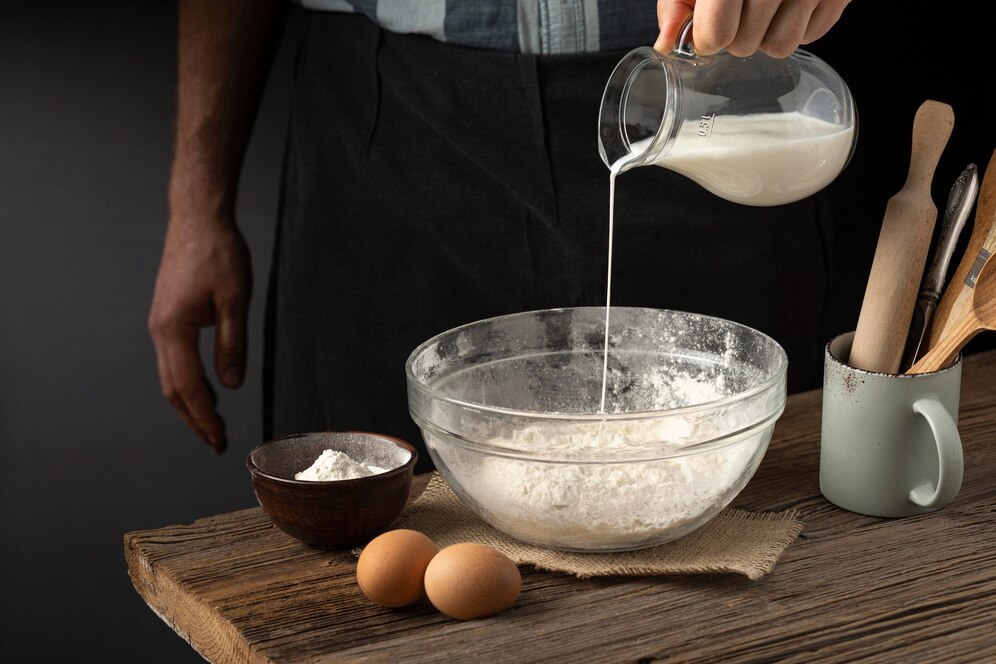 A person pours milk into a glass bowl of flour, surrounded by eggs, a bowl of flour, and kitchen utensils on a wooden table.