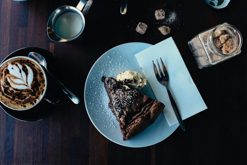 A slice of rich chocolate cake topped with ice cream and powdered sugar, accompanied by latte art coffee and sugar cubes on a wooden table.