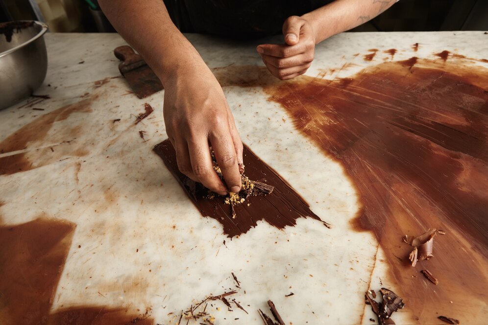 A hand breaking chocolate over a marble surface, surrounded by shavings and cocoa remnants, capturing the art of chocolate making.