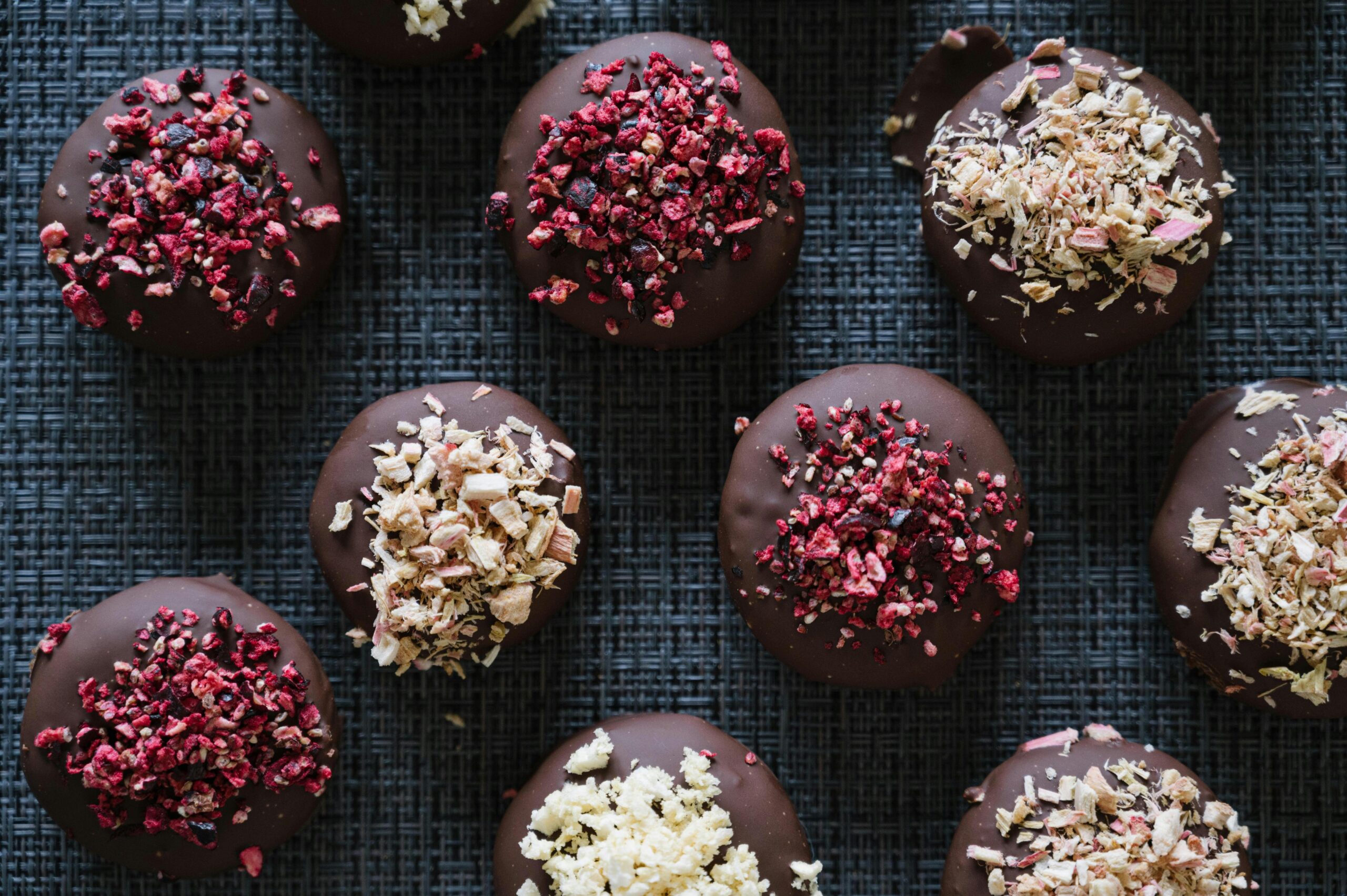 An assortment of chocolate-dipped cookies topped with crushed raspberries and oats, arranged on a textured dark surface.