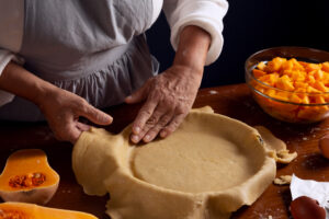 A person rolls out dough for a pie crust on a wooden surface, surrounded by fresh pumpkin pieces and other baking ingredients.