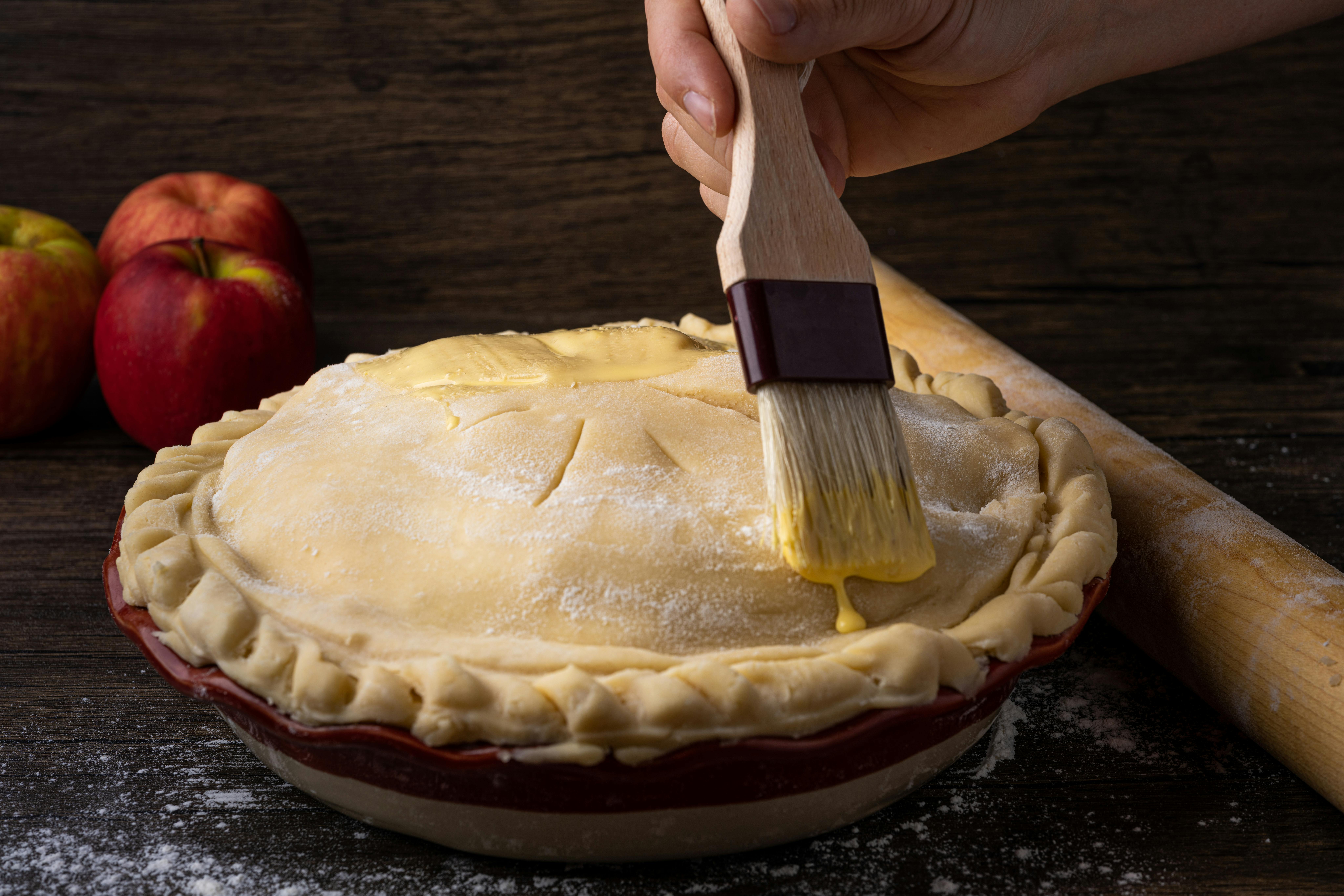A hand brushes egg wash onto an unbaked apple pie, with red apples and a rolling pin in the background on a wooden surface.