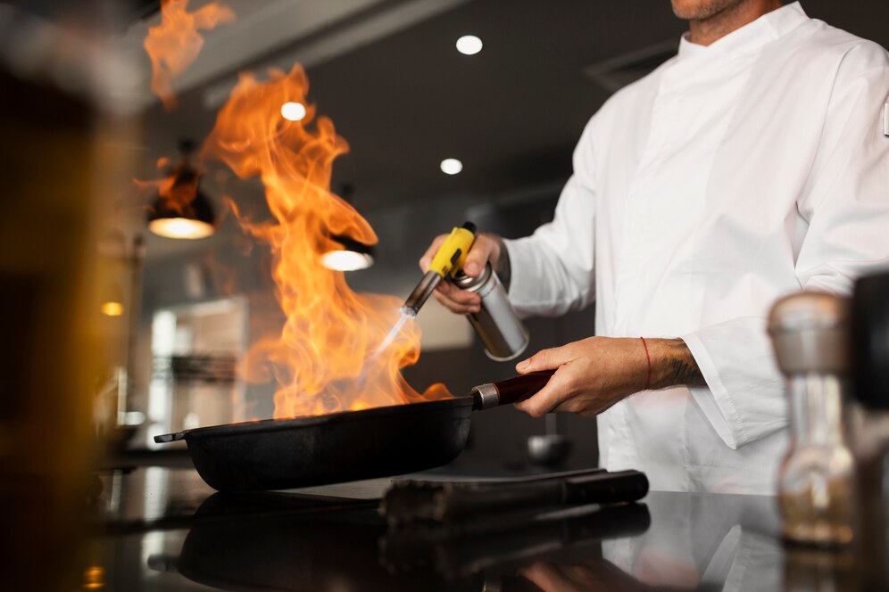 A chef in a white uniform uses a kitchen torch to ignite flames in a frying pan, showcasing culinary skill in a modern kitchen.