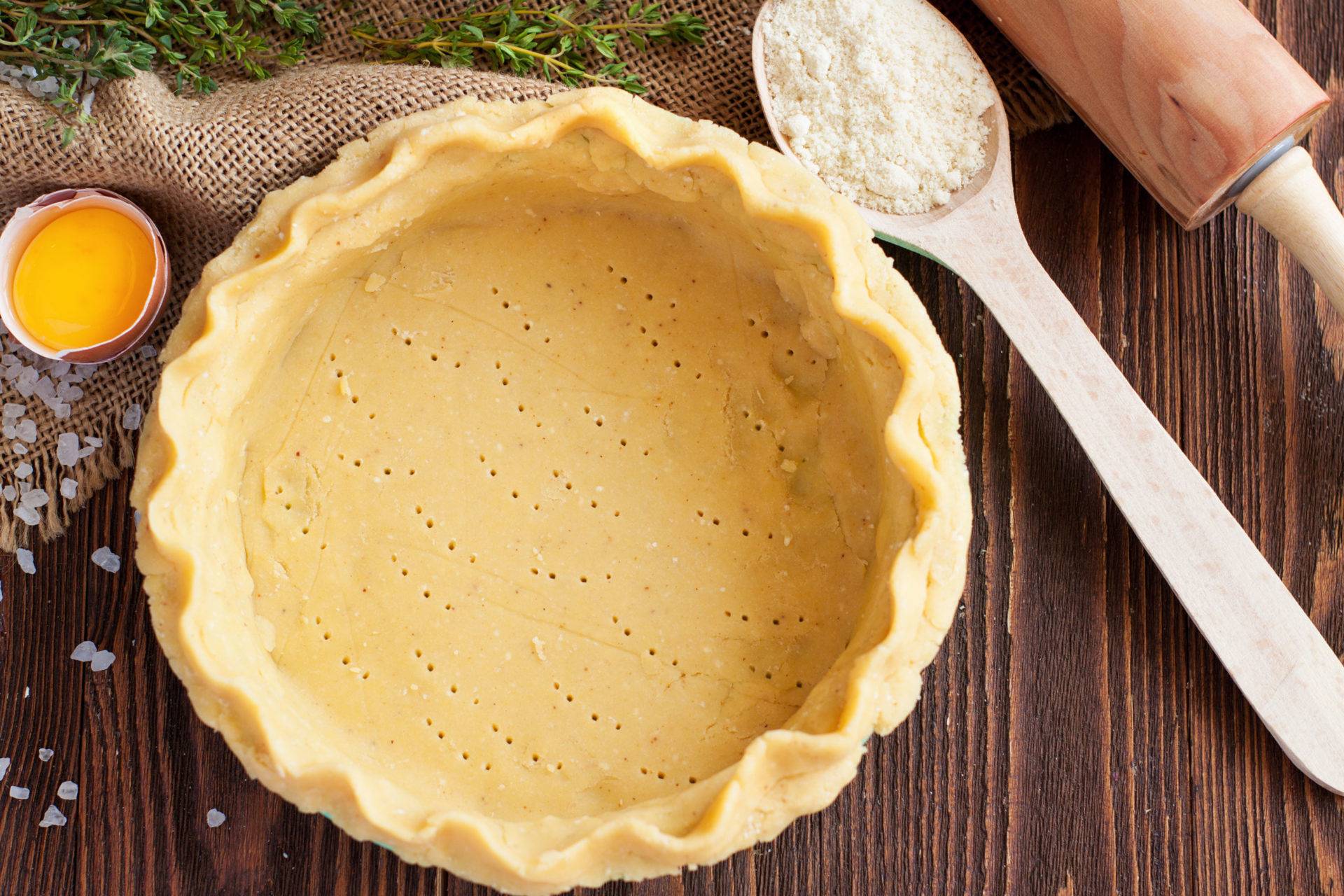A prepared pie crust sits on a wooden surface, accompanied by an egg yolk, flour, and fresh thyme, ready for baking.