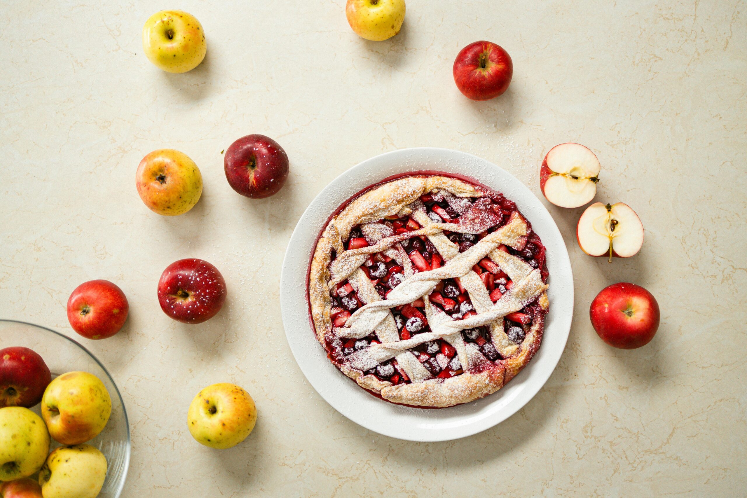 A freshly baked apple pie on a white plate, surrounded by assorted whole and sliced apples on a light marble surface.