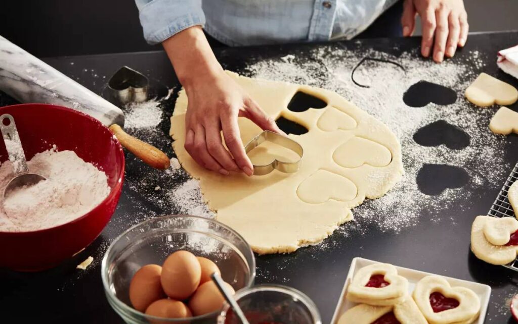 A person uses a heart-shaped cookie cutter on rolled dough, surrounded by flour, eggs, and baking tools on a dark countertop.