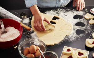 A person uses a heart-shaped cookie cutter on rolled dough, surrounded by flour, eggs, and baking tools on a dark countertop.