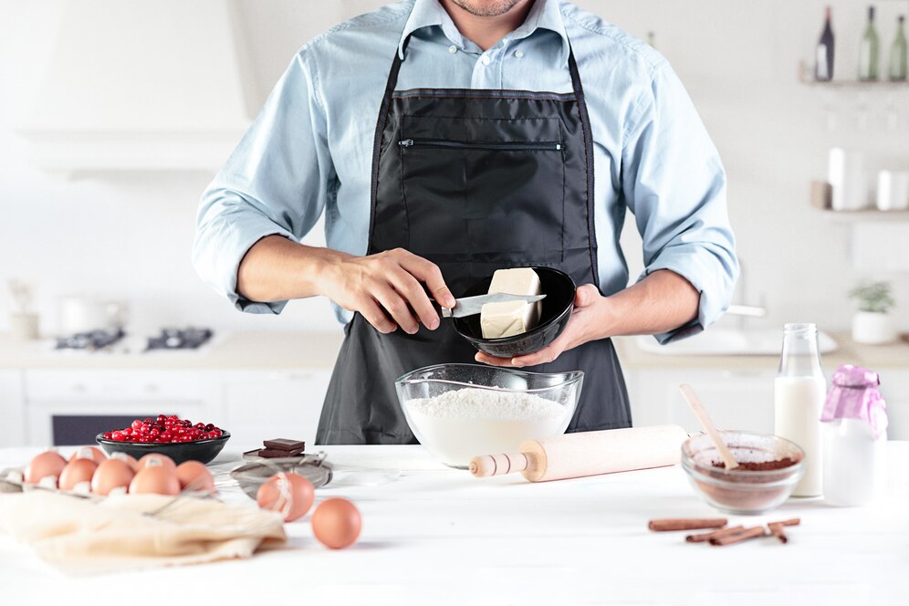 A man in an apron prepares ingredients, holding butter over a bowl of flour with eggs, chocolate, and milk on the counter.