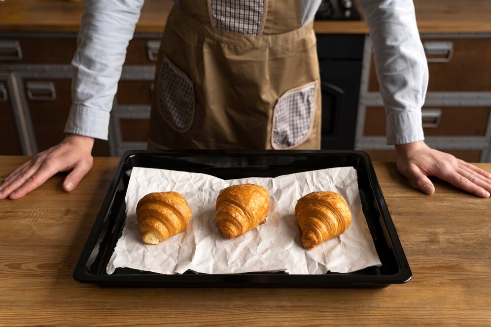 A person in a brown apron stands over a tray with three golden croissants on parchment paper, resting on a wooden table.