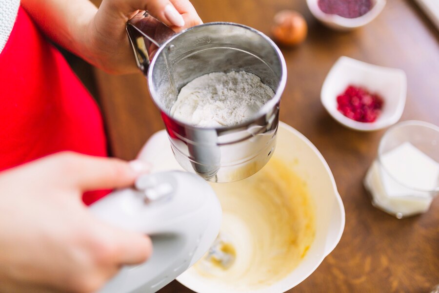 A person sifts flour into a mixing bowl with a handheld mixer nearby, surrounded by ingredients on a wooden countertop.