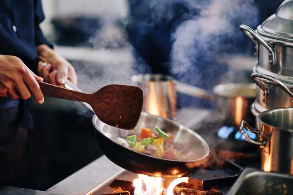 A chef stirs colorful vegetables in a smoky frying pan over an open flame in a bustling kitchen. Steam rises around them.