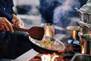 A chef stirs colorful vegetables in a smoky frying pan over an open flame in a bustling kitchen. Steam rises around them.