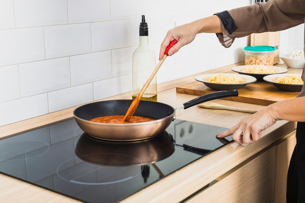 A person stirs tomato sauce in a pan on an induction cooktop, with plates of pasta in the background and olive oil bottle nearby.