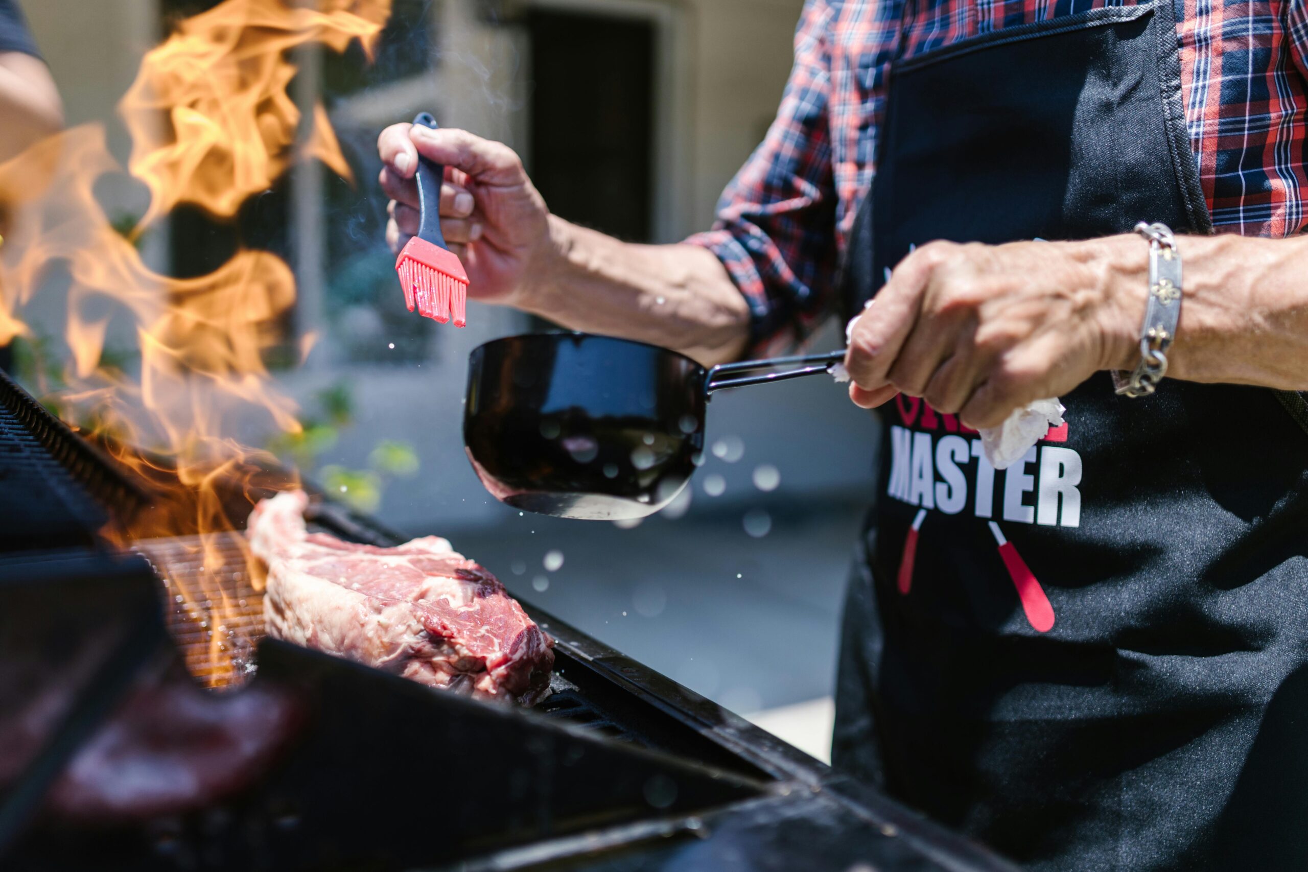 A person wearing a grill master apron brushes barbecue sauce onto sizzling meat over a flame-grilled barbecue.
