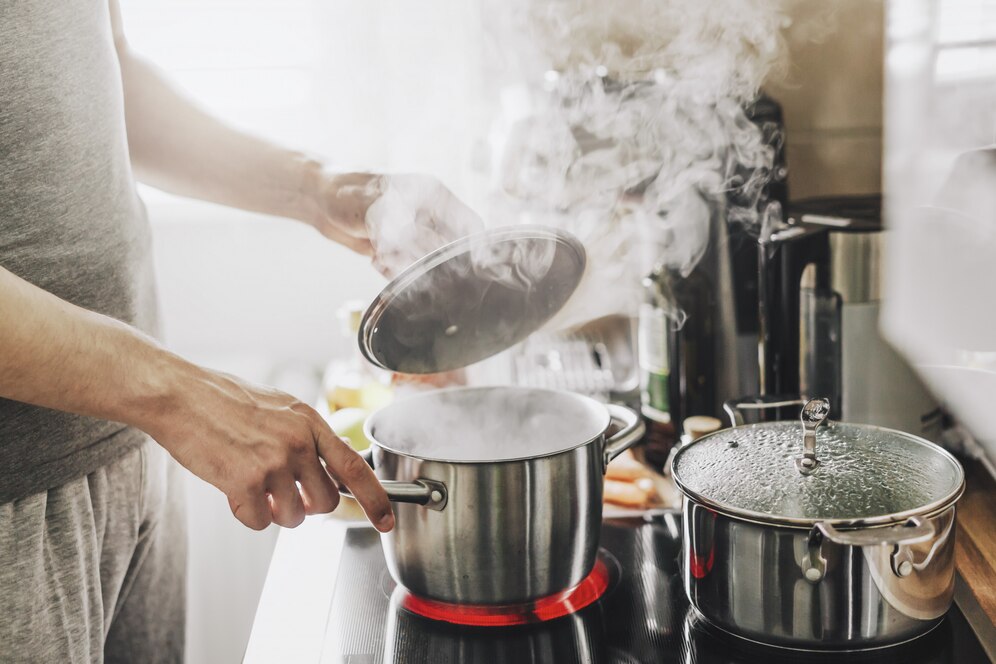 A person in a gray shirt lifts the lid off a steaming pot on a stovetop, with another pot nearby.