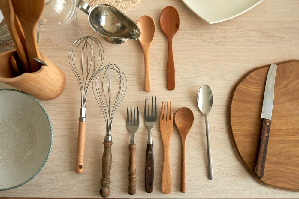 A collection of kitchen utensils, including whisks, forks, spoons, and a knife, arranged on a wooden surface with bowls and a plate.