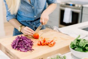 A person in a denim shirt slices a tomato on a wooden cutting board, surrounded by chopped red cabbage and fresh greens.