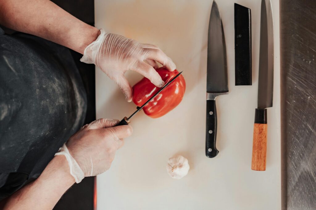 A chef in gloves slices a red pepper on a cutting board, surrounded by knives and a garlic bulb.