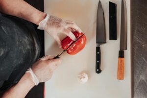 A chef in gloves slices a red pepper on a cutting board, surrounded by knives and a garlic bulb.