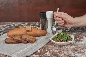A hand holds a spoon above a bowl of lentils garnished with herbs, beside slices of bread and salt and pepper shakers.