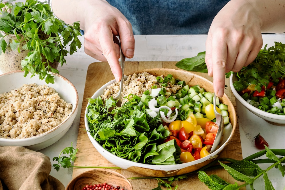 A person mixes a colorful salad with leafy greens, cherry tomatoes, and cucumbers in a wooden bowl, surrounded by fresh ingredients.