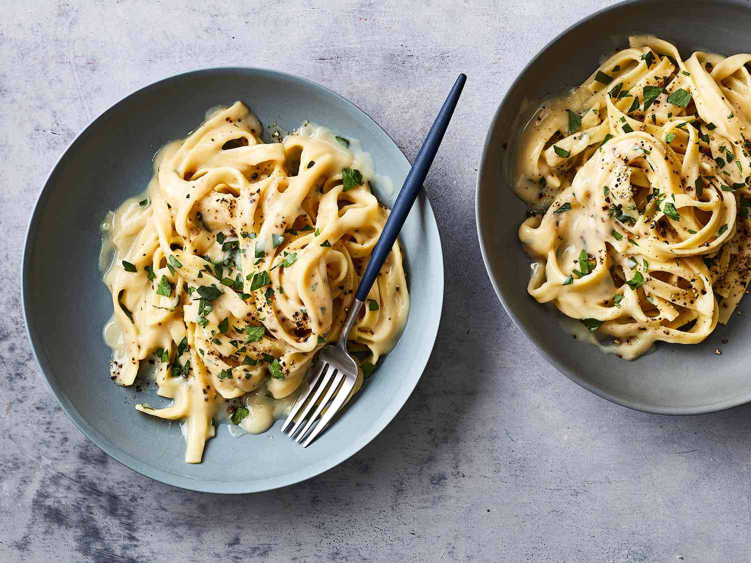 Two bowls of creamy fettuccine pasta garnished with fresh herbs and black pepper, with a fork resting in one bowl.