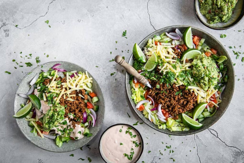 Two vibrant salad bowls filled with seasoned meat, cheese, fresh veggies, guacamole, and lime wedges, accompanied by a creamy dip.