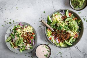 Two vibrant salad bowls filled with seasoned meat, cheese, fresh veggies, guacamole, and lime wedges, accompanied by a creamy dip.