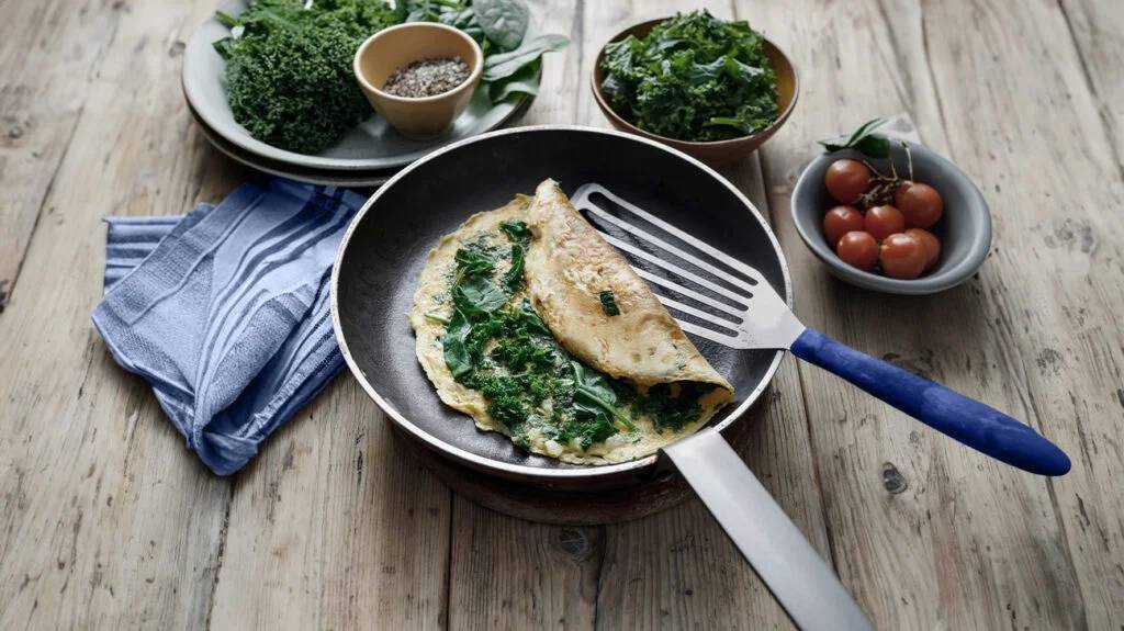 A spinach-filled omelette in a frying pan, surrounded by fresh greens, tomatoes, and a bowl of seasonings on a rustic wooden table.
