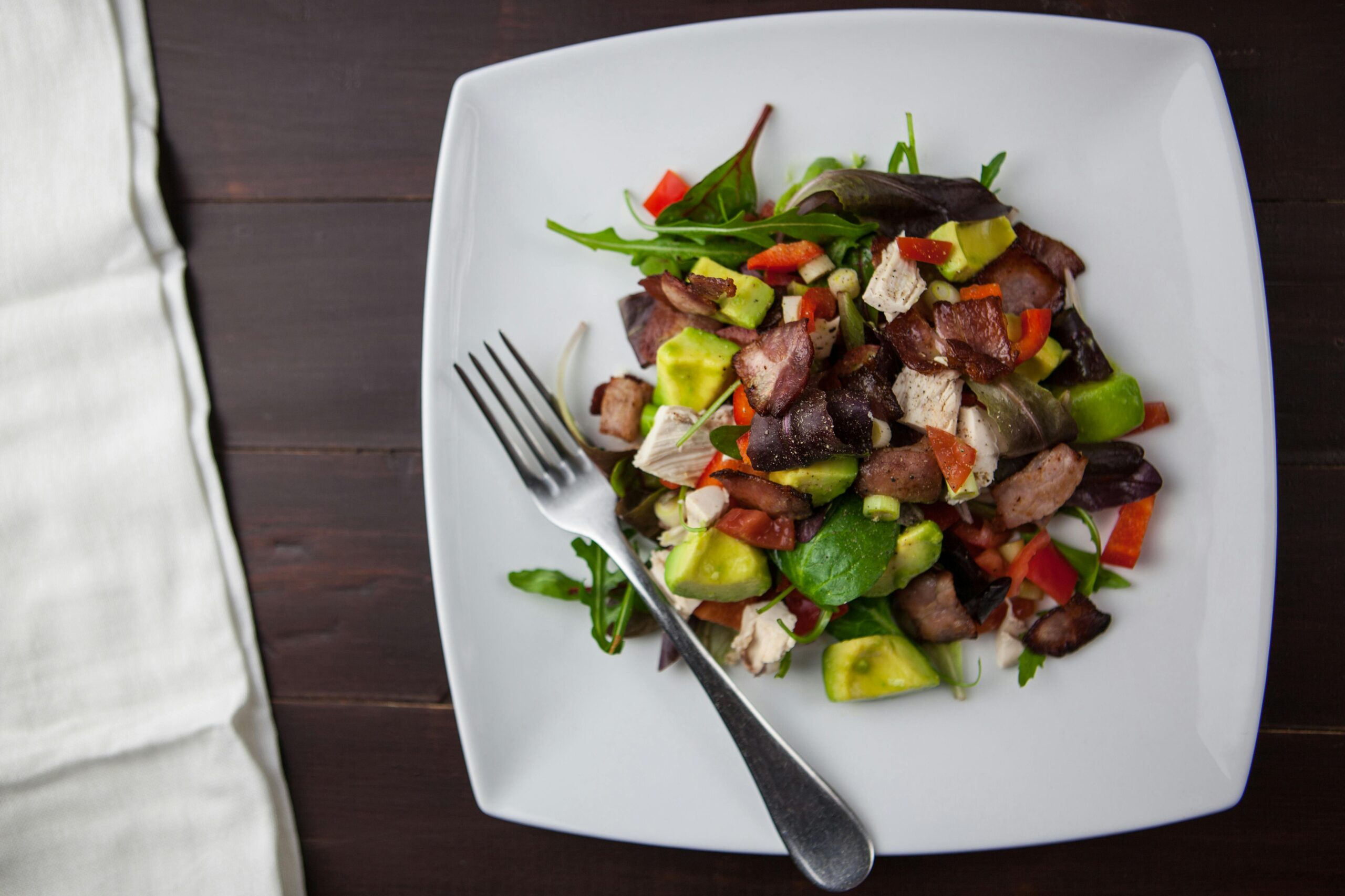 A vibrant salad topped with greens, diced chicken, bacon, red and green peppers, and avocado, served on a white plate.