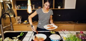 A person in a striped shirt cooks in a modern kitchen, flipping fried fillets and preparing plates with fresh ingredients.