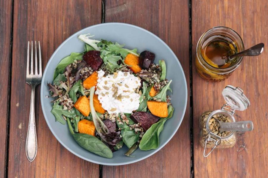 A colorful salad with spinach, quinoa, roasted sweet potatoes, beets, and a dollop of yogurt, alongside olive oil and seeds.