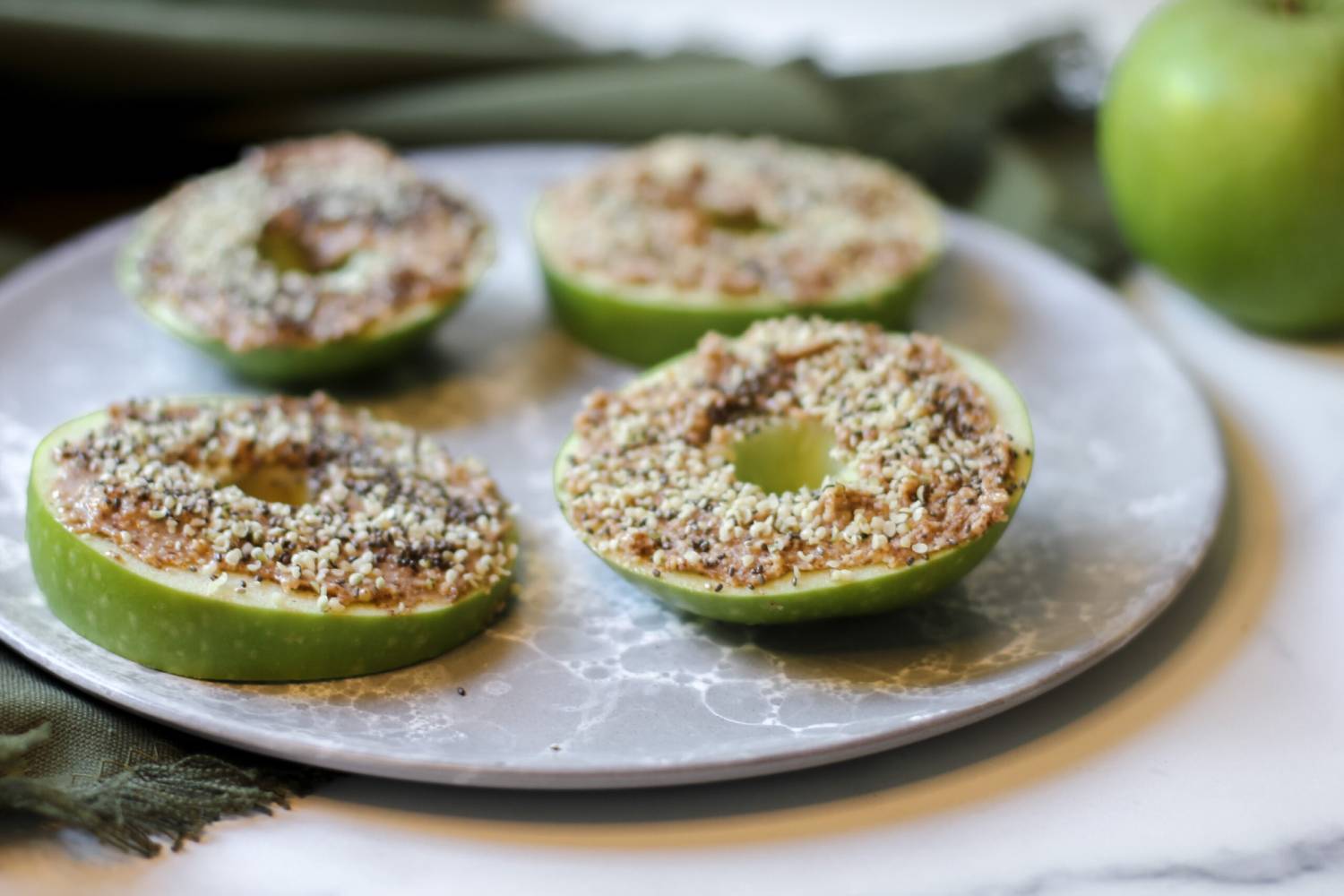 Four halved green apples topped with a crumbly mixture on a marble plate, with a whole apple and green cloth in the background.