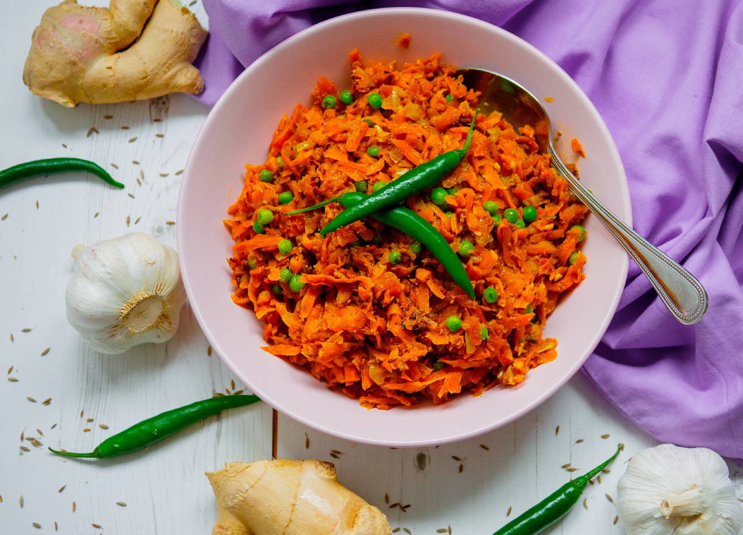 A vibrant bowl of shredded carrot dish with green peas, topped with green chilies, surrounded by garlic and ginger on a white table.