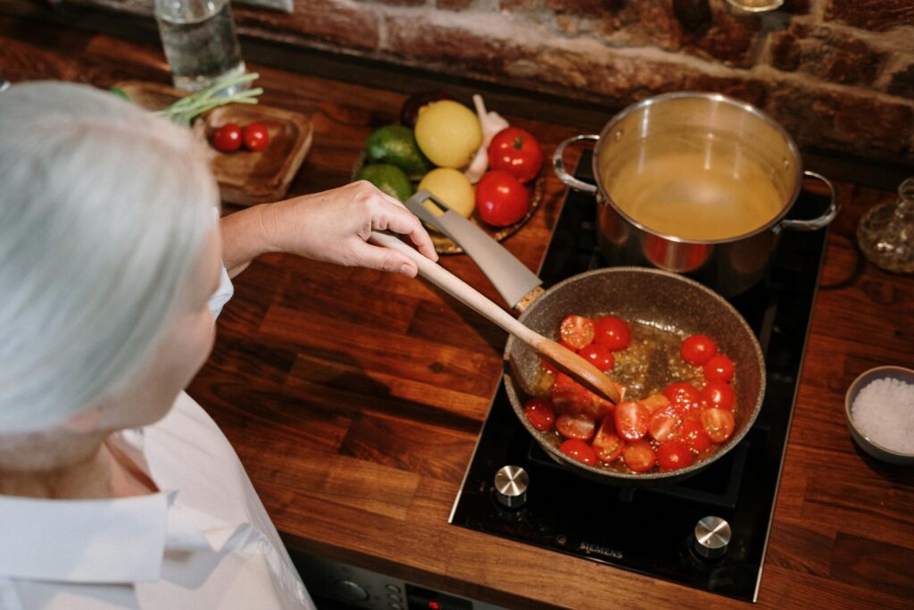A woman with gray hair stirs cherry tomatoes in a frying pan on a wooden countertop, surrounded by fresh ingredients and boiling pasta.