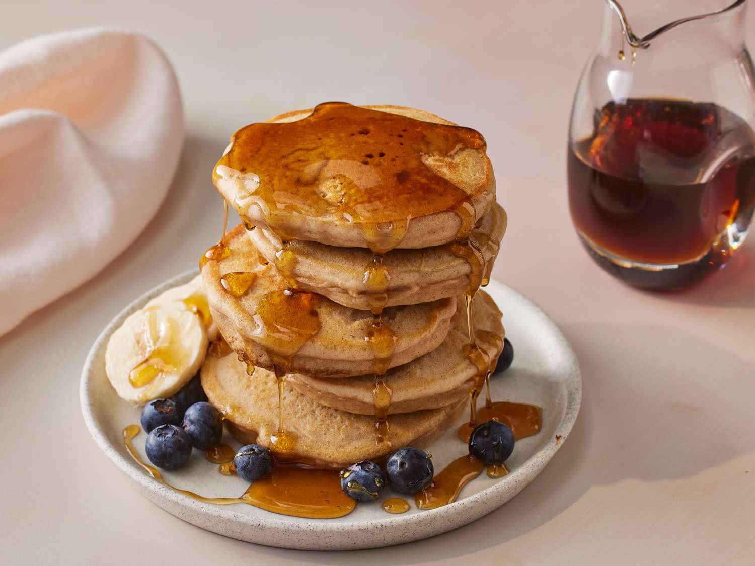 A stack of fluffy pancakes drizzled with syrup, topped with fresh blueberries and a banana slice, beside a pitcher of maple syrup.