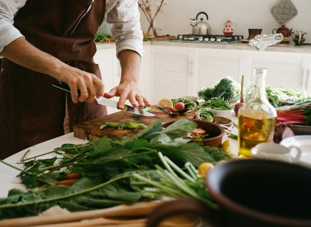 A person chops fresh vegetables on a wooden board, surrounded by herbs, olive oil, and an array of colorful ingredients in a bright kitchen.