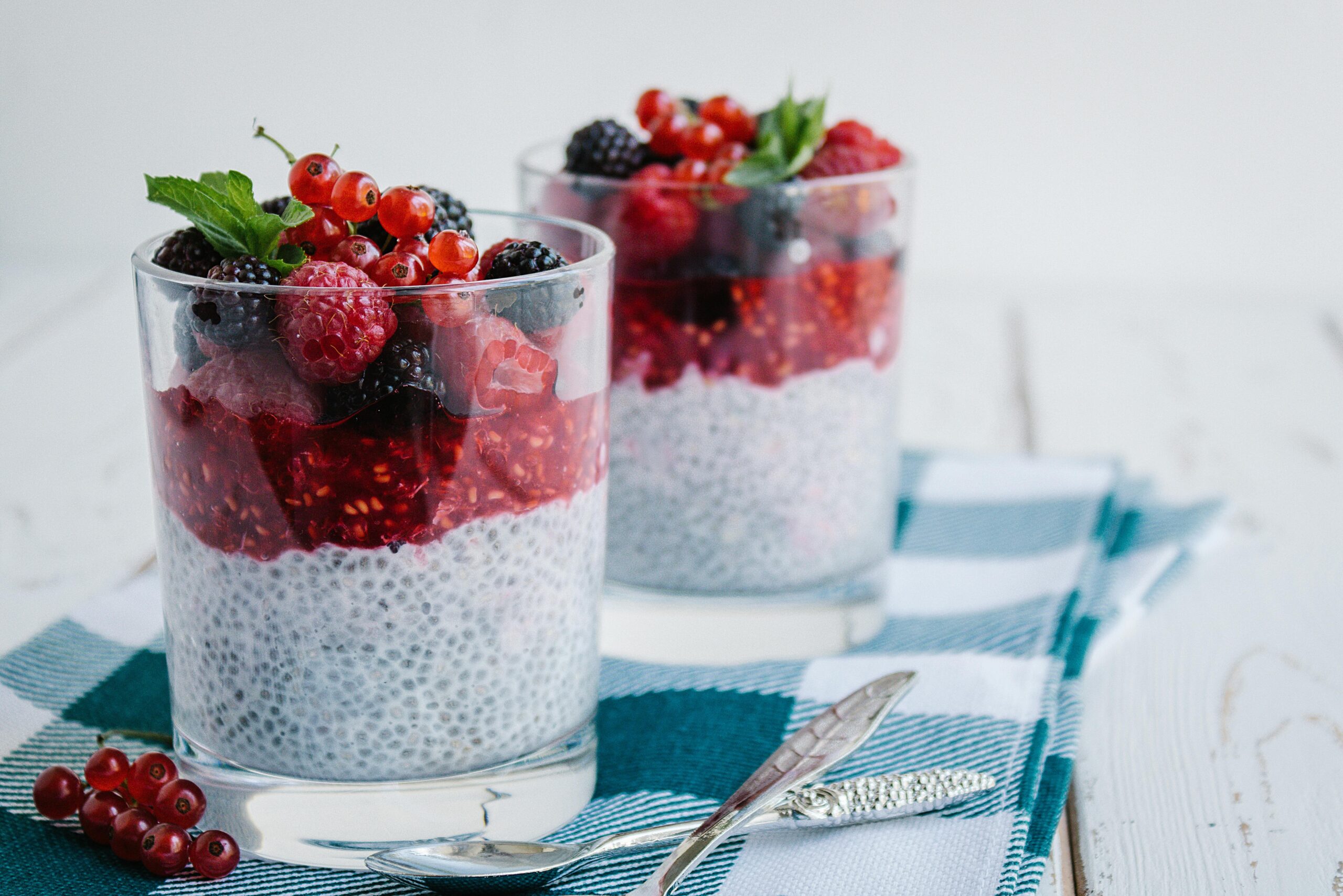 Two glass cups of layered chia pudding topped with mixed berries and a sprig of mint, served on a blue checkered napkin.