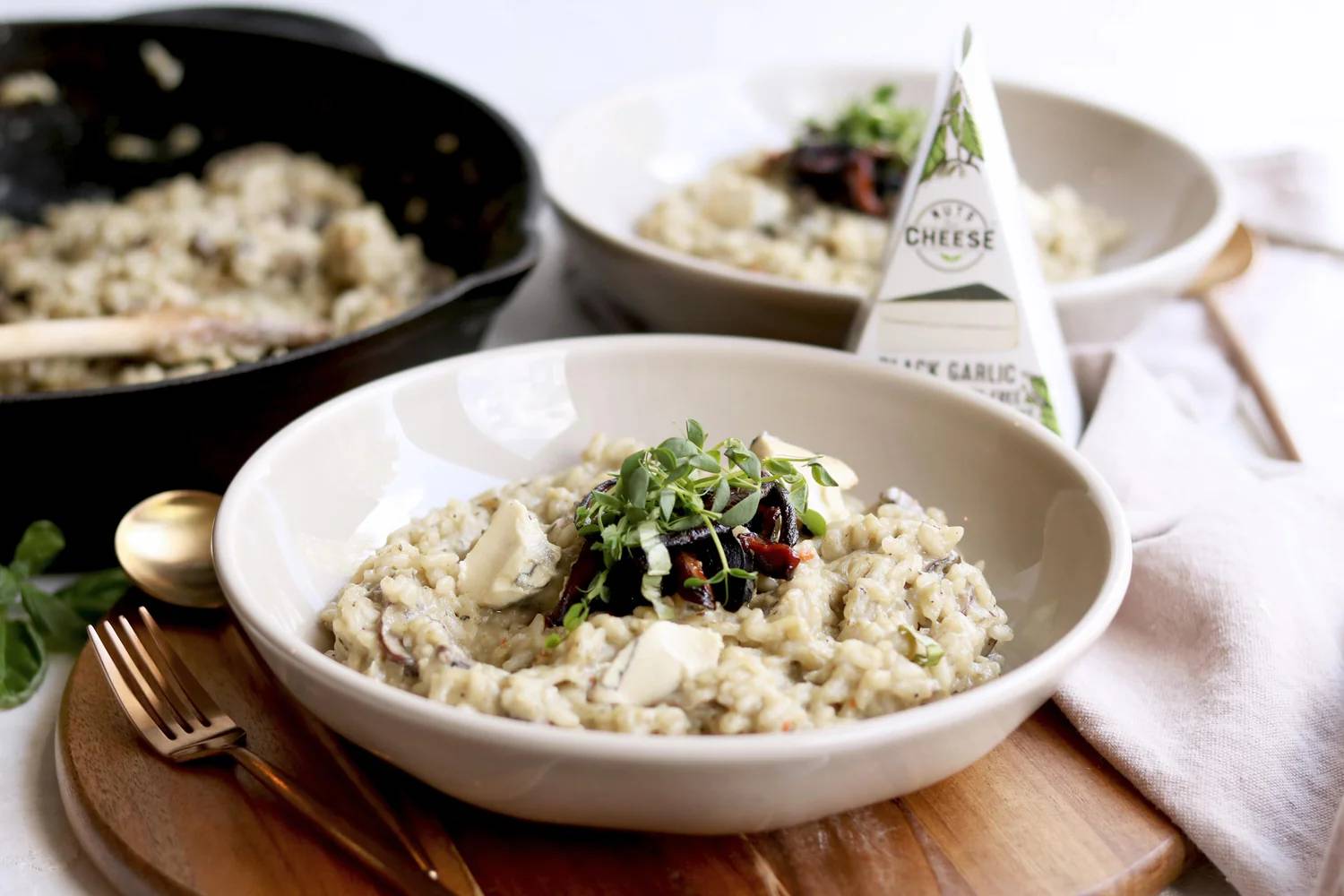 Creamy risotto topped with fresh herbs and black garlic, served in a white bowl on a wooden board with a spoon and fork.