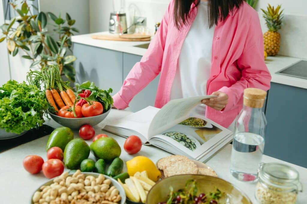A person in a pink shirt reads a cookbook amidst a colorful array of fresh vegetables, fruits, and snacks on a kitchen counter.